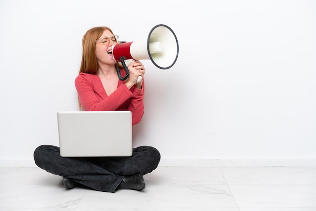 Young redhead woman with a laptop sitting on the floor isolated on white background shouting through a megaphone