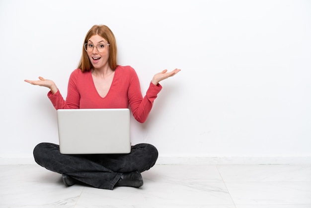 Young redhead woman with a laptop sitting on the floor isolated on white background with shocked facial expression