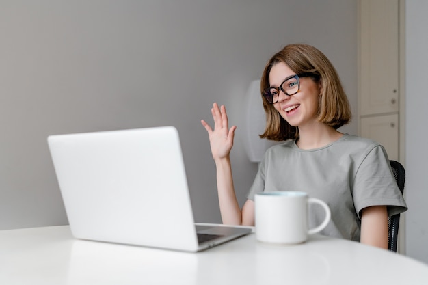 Young relaxed caucasian woman smiling waving with laptop in the apartment with cup on the table