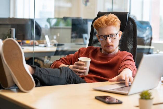 Young relaxed office worker in casualwear having glass of coffee at break while sitting in armchair with his legs on table in front of laptop