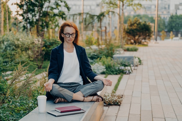 Young relaxed redhead woman meditates in park feels calm sits in lotus pose outdoor