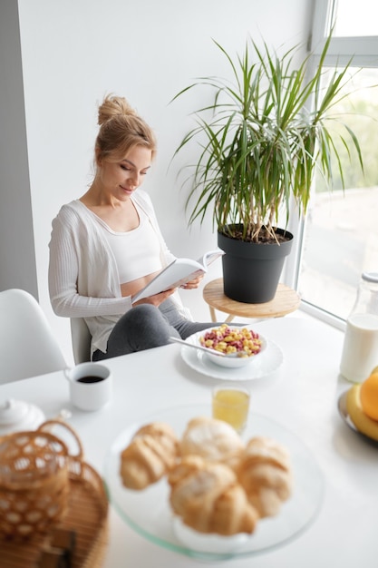 Young relaxed woman reading after sitting by kitchen table after healthy breakfast at home in morning