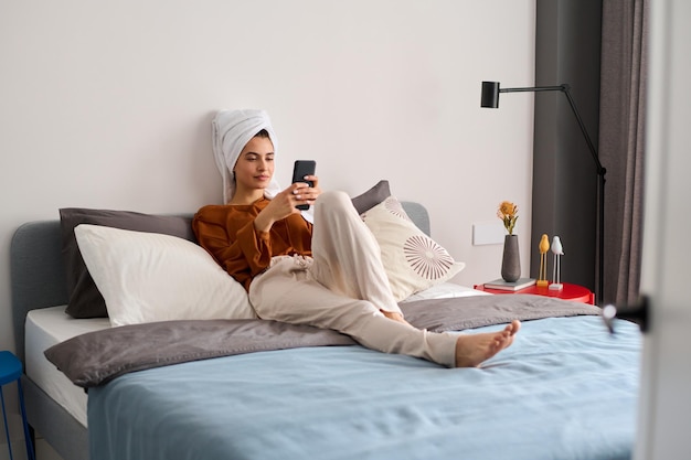 Young restful female in home wear sitting on double bed and texting