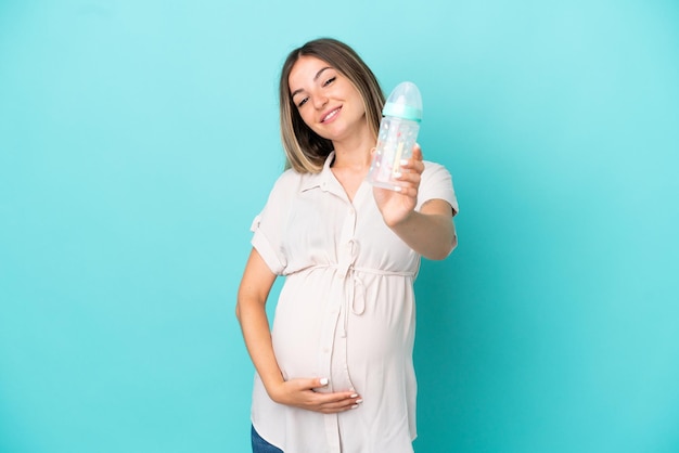 Young Romanian woman isolated on blue background pregnant and holding a feeding bottle