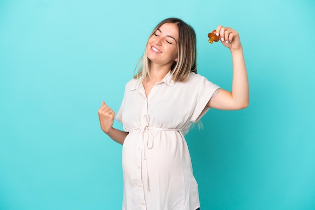 Young Romanian woman isolated on blue background pregnant and holding a pacifier and celebrating a victory