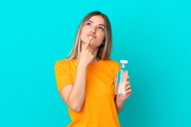 Young Romanian woman with a bottle of water isolated on blue background having doubts while looking up