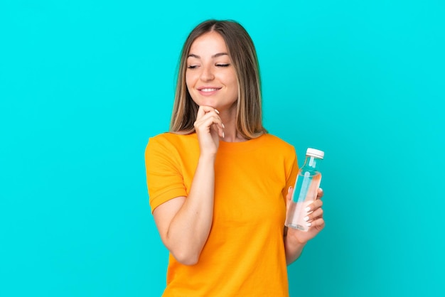 Young Romanian woman with a bottle of water isolated on blue background looking to the side and smiling