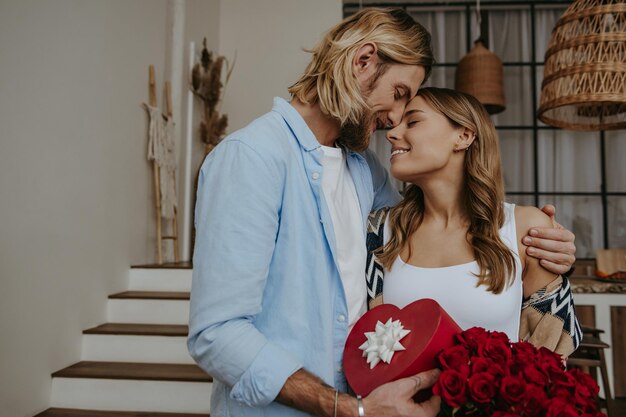 Young romantic couple embracing and holding gift box and flowers while celebrating anniversary