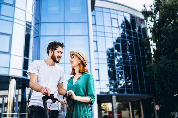 A young romantic couple with electric scooters on a date, walking in the city. Young woman in hat and man enjoy a walk