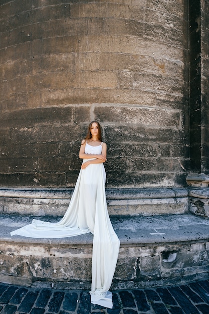 Young romantic elegant girl in long white dress posing over stone ancient wall