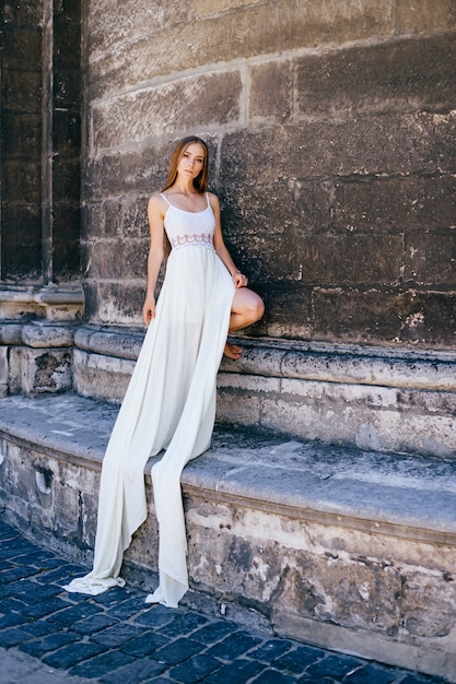 Young romantic elegant girl in long white dress posing over stone ancient wall