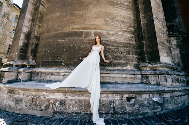 Young romantic elegant girl in long white dress posing over stone ancient wall