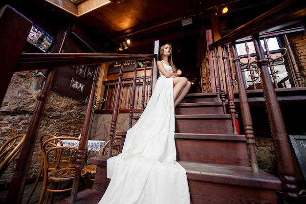 Young romantic elegant girl in long white flowy dress posing on the stairs indoor