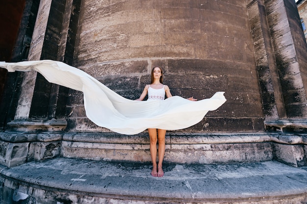 Young romantic elegant girl in long white flowy dress posing over stone ancient wall