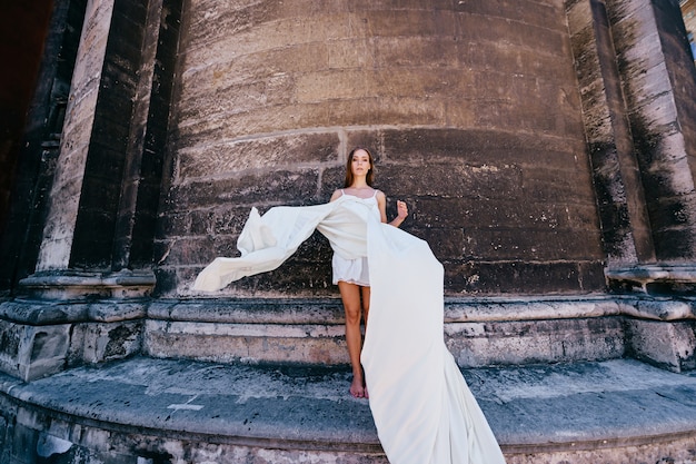 Young romantic elegant girl in long white flowy dress posing over stone ancient wall