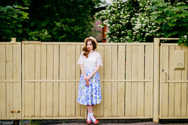 Young russian beauty. Attractive girl in vintage retro long skirt, white old fashioned top and curly red hair and straw hat posing for camera with fence and green trees