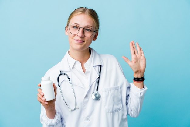 Young russian doctor woman holding pills bottle on blue smiling cheerful showing number five with fingers.