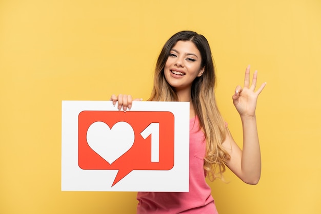 Young Russian girl isolated on yellow background holding a placard with Like icon and celebrating a victory