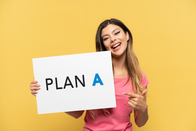 Young Russian girl isolated on yellow background holding a placard with the message PLAN A and  pointing it