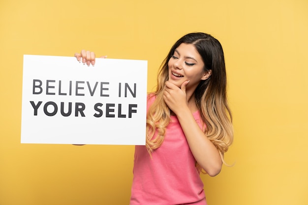 Young Russian girl isolated on yellow background holding a placard with text Believe In Your Self and thinking