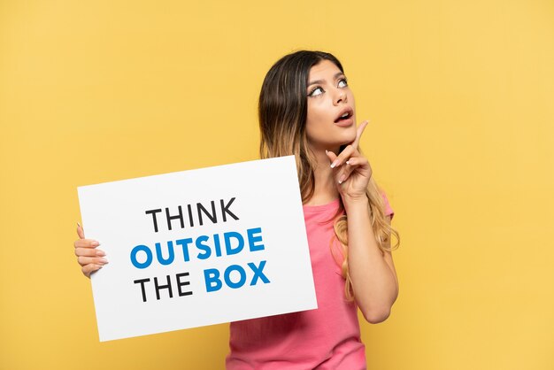 Young Russian girl isolated on yellow background holding a placard with text Think Outside The Box and thinking