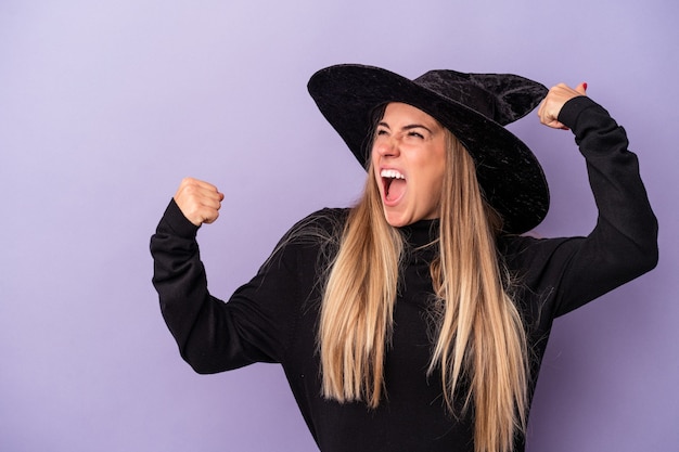 Young Russian woman disguised as a witch celebrating Halloween isolated on purple background raising fist after a victory, winner concept.