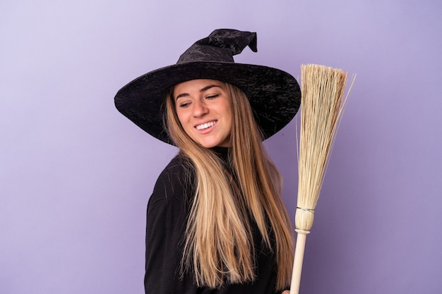 Young Russian woman disguised as a witch holding a broom isolated on purple background looks aside smiling, cheerful and pleasant.