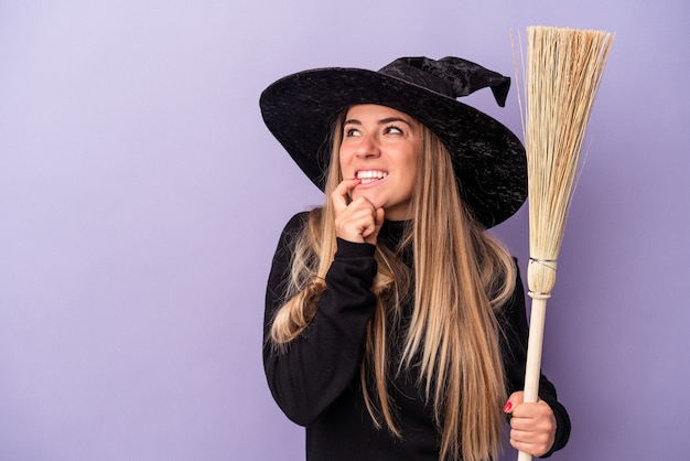 Young Russian woman disguised as a witch holding a broom isolated on purple background relaxed thinking about something looking at a copy space.