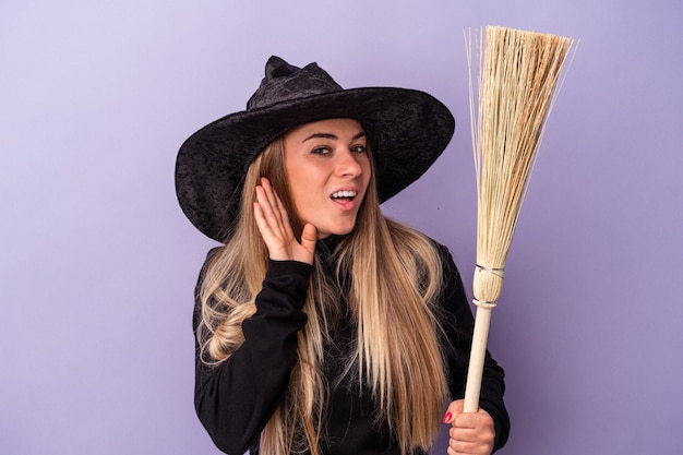 Young Russian woman disguised as a witch holding a broom isolated on purple background trying to listening a gossip.