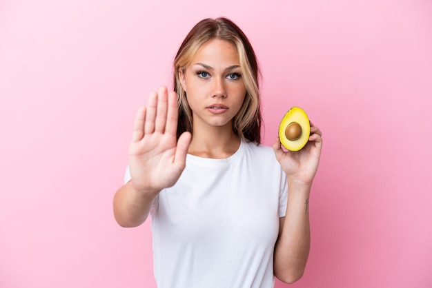 Young Russian woman holding avocado isolated on pink background making stop gesture