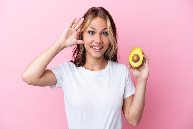 Young Russian woman holding avocado isolated on pink background with surprise expression