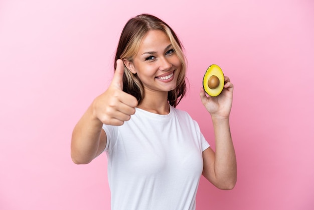 Young Russian woman holding avocado isolated on pink background with thumbs up because something good has happened