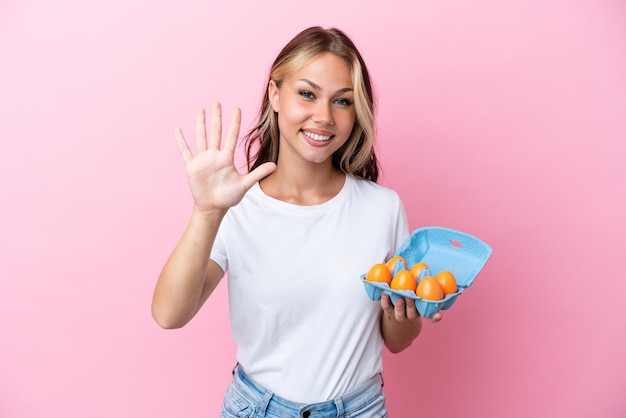 Young Russian woman holding eggs isolated on pink background counting five with fingers