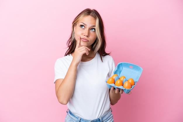 Young Russian woman holding eggs isolated on pink background having doubts while looking up