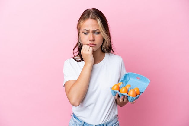 Young Russian woman holding eggs isolated on pink background having doubts