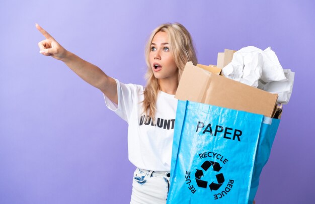 Young Russian woman holding a recycling bag full of paper to recycle isolated on purple wall pointing away