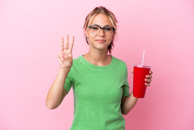 Young Russian woman holding a refreshment isolated on pink background happy and counting three with fingers