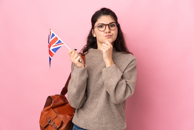 Young Russian woman holding an United Kingdom flag isolated on pink background having doubts and thinking