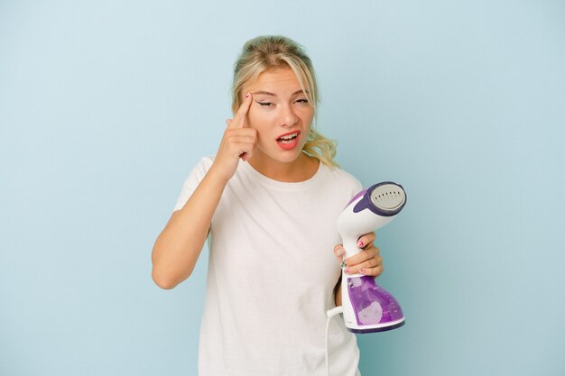 Young Russian woman holding vertical iron isolated on blue background showing a disappointment gesture with forefinger.