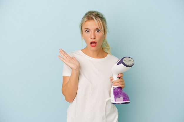 Young Russian woman holding vertical iron isolated on blue background surprised and shocked.