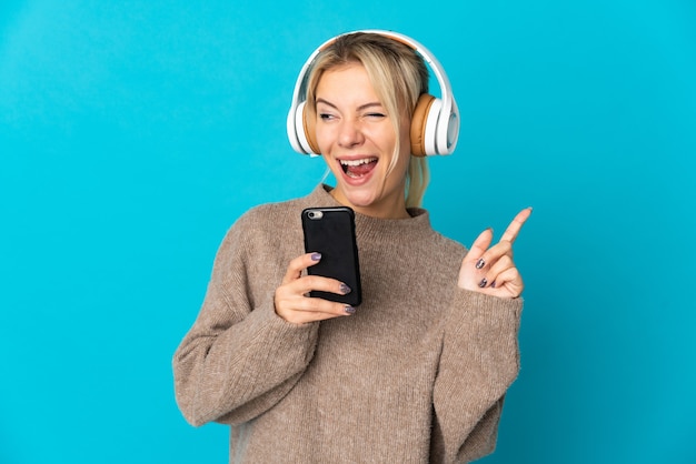 Young Russian woman isolated on blue listening music with a mobile and singing