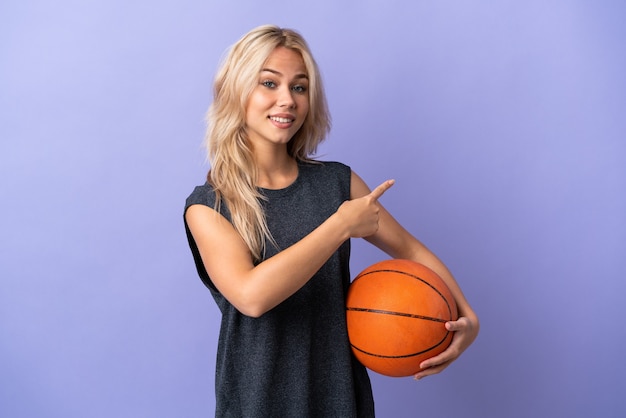 Young Russian woman isolated on purple wall playing basketball and pointing to the lateral