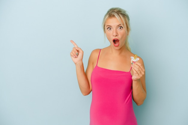 Young Russian woman pregnant holding a pacifier isolated on blue background pointing to the side