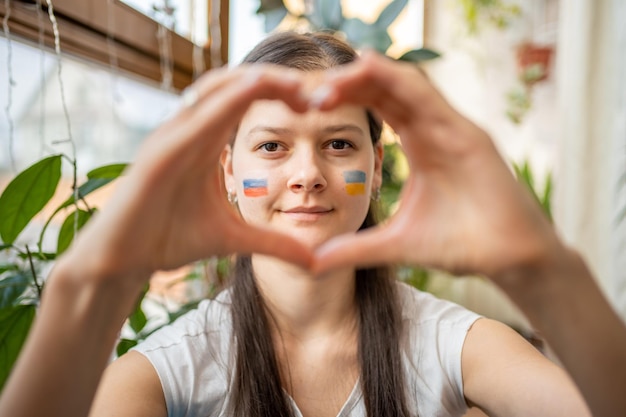 Young russianukrainian girl with the flag of ukraine and russia on her face is showing heart by hand