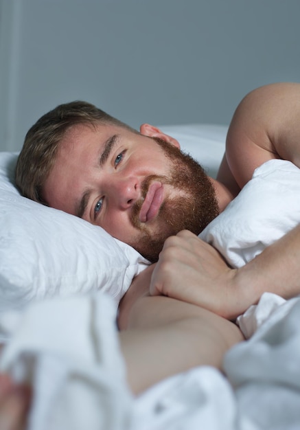 Young sad depressed unhappy worried man in the bed in bedroom at home in the morning lying under white blanket suffering from insomnia problems with sleep at night