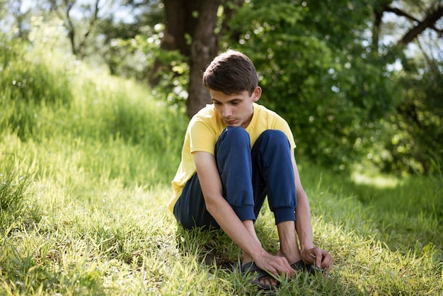 Young sad guy sitting on the grass alone in the forest during the day