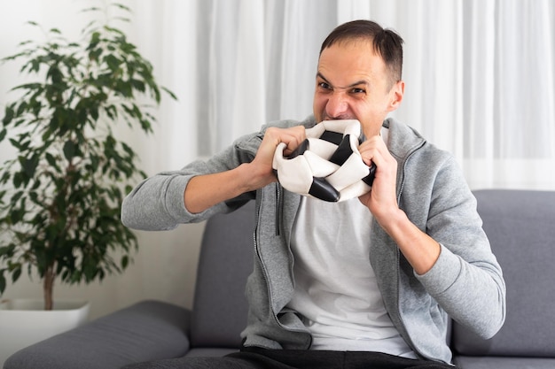 Young sad unhappy man fan in brown shirt hold remote controller spread hands cheer up support football team sit on blue sofa with soccer ball rest watch tv indoors room gray wall Sport leisure concept