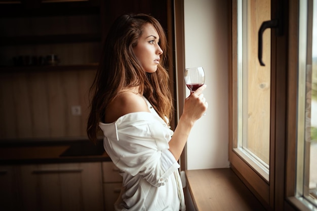 Young sad woman is drinking red wine and looking through the window, feels unhappy and depressed, suffering from abuse, harassment, or depression or heartbreak.