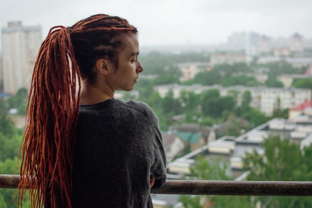 Young sad woman with red dreadlocks looks at the city from the balcony after the rain and waiting.