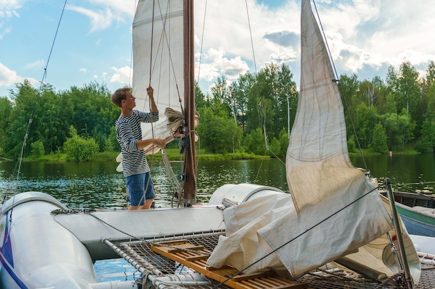 Young sailor raises a sail on a small sailing catamaran moored in a wooded bay